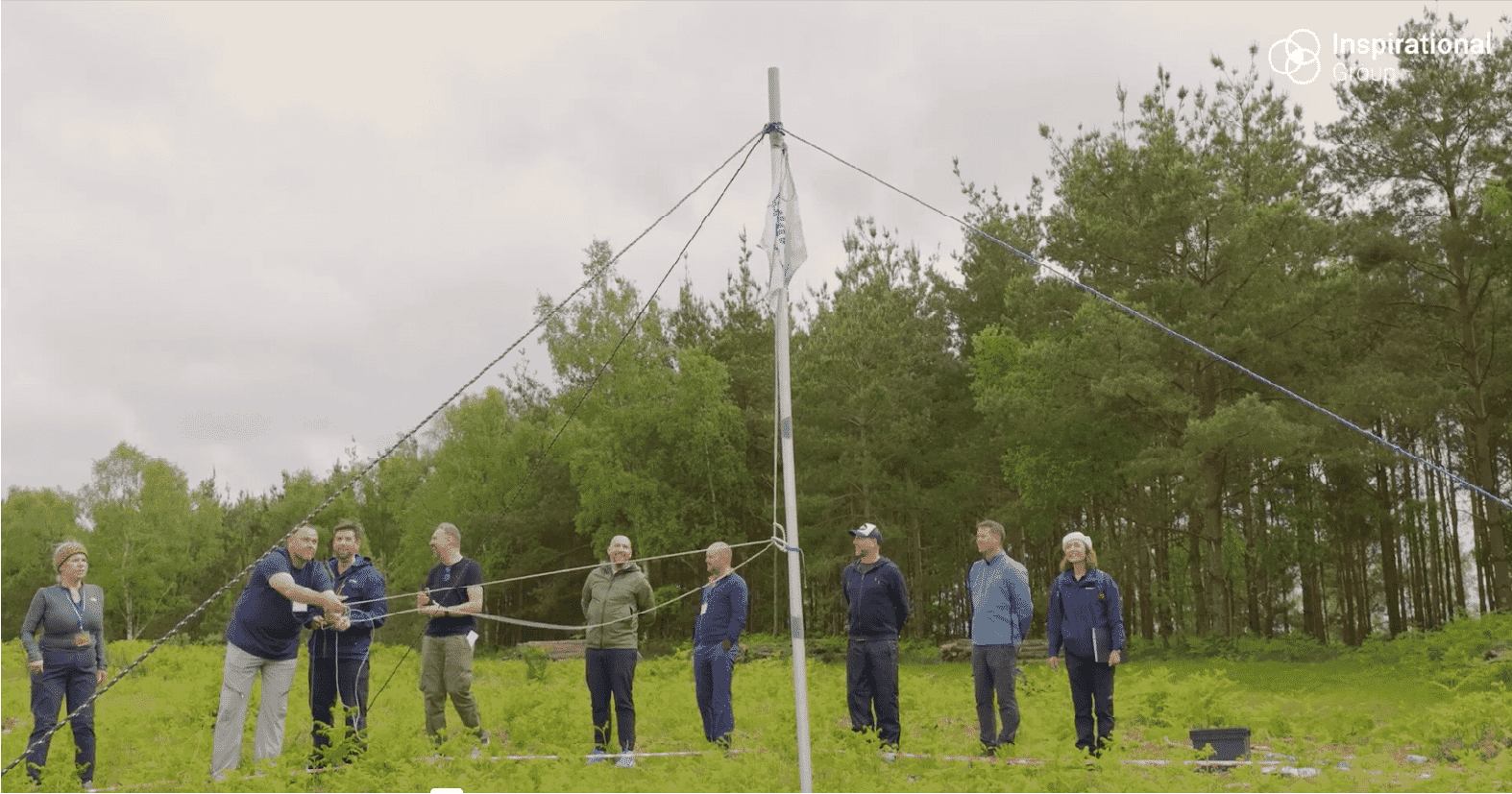 “A team of participants from Bourne Leisure collaborate during an outdoor leadership challenge, pulling ropes to stabilise a tall pole in a woodland setting.”