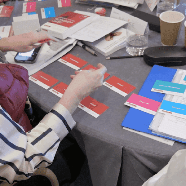 Participant’s hands sorting red and blue prompt cards on a workshop table, surrounded by notebooks, stationery, and training materials during an interactive group exercise.