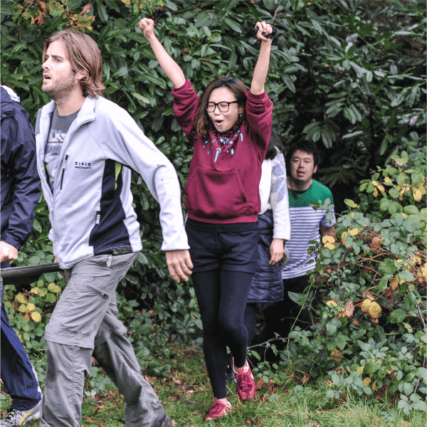 “A participant raises their arms in celebration after completing an outdoor team challenge, with other participants walking along a wooded path.”